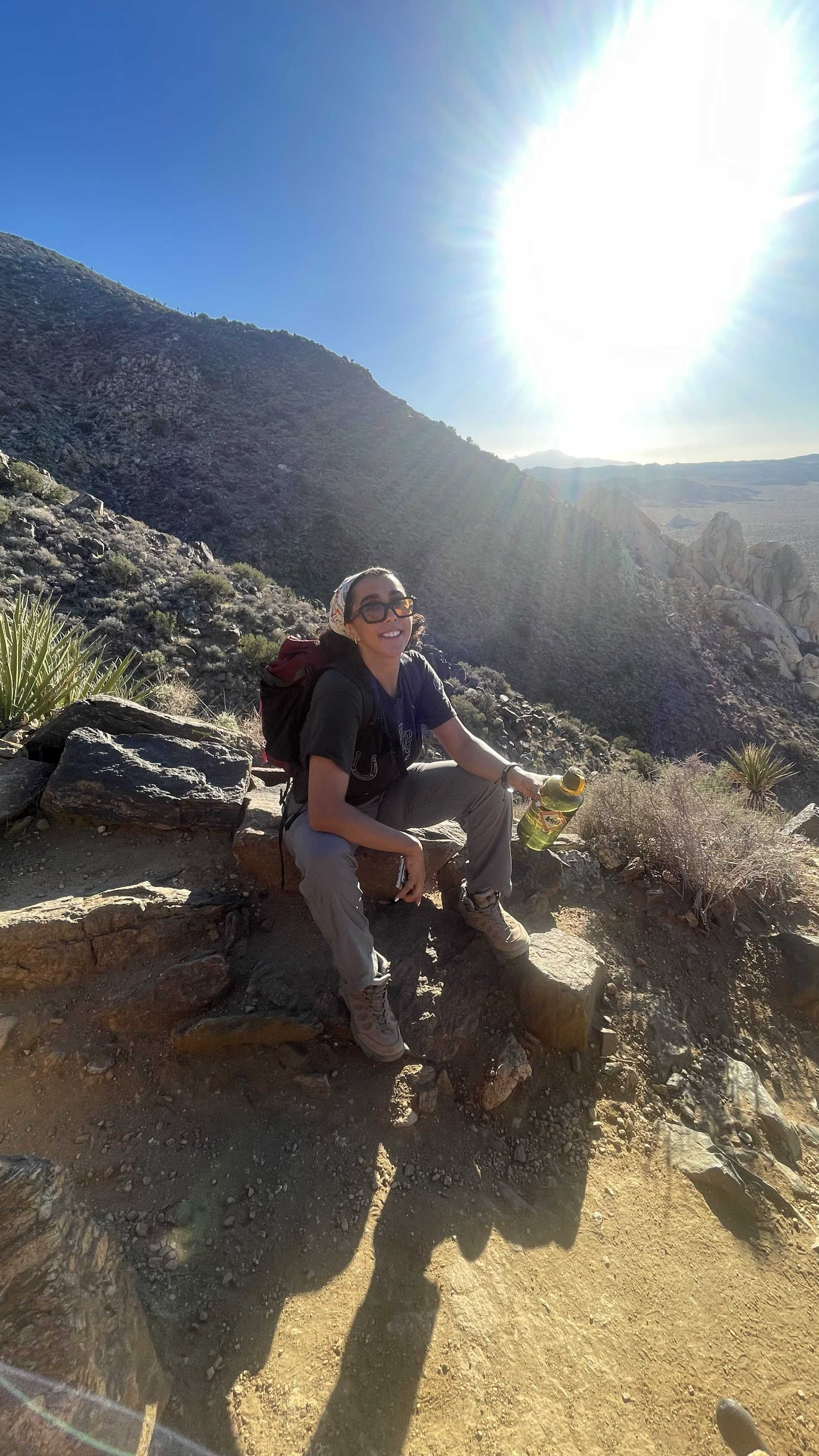 Aysheh Alrfooh sits on a mountain rock in her hiking outfit, sunlight glowing behind her like a spotlight.