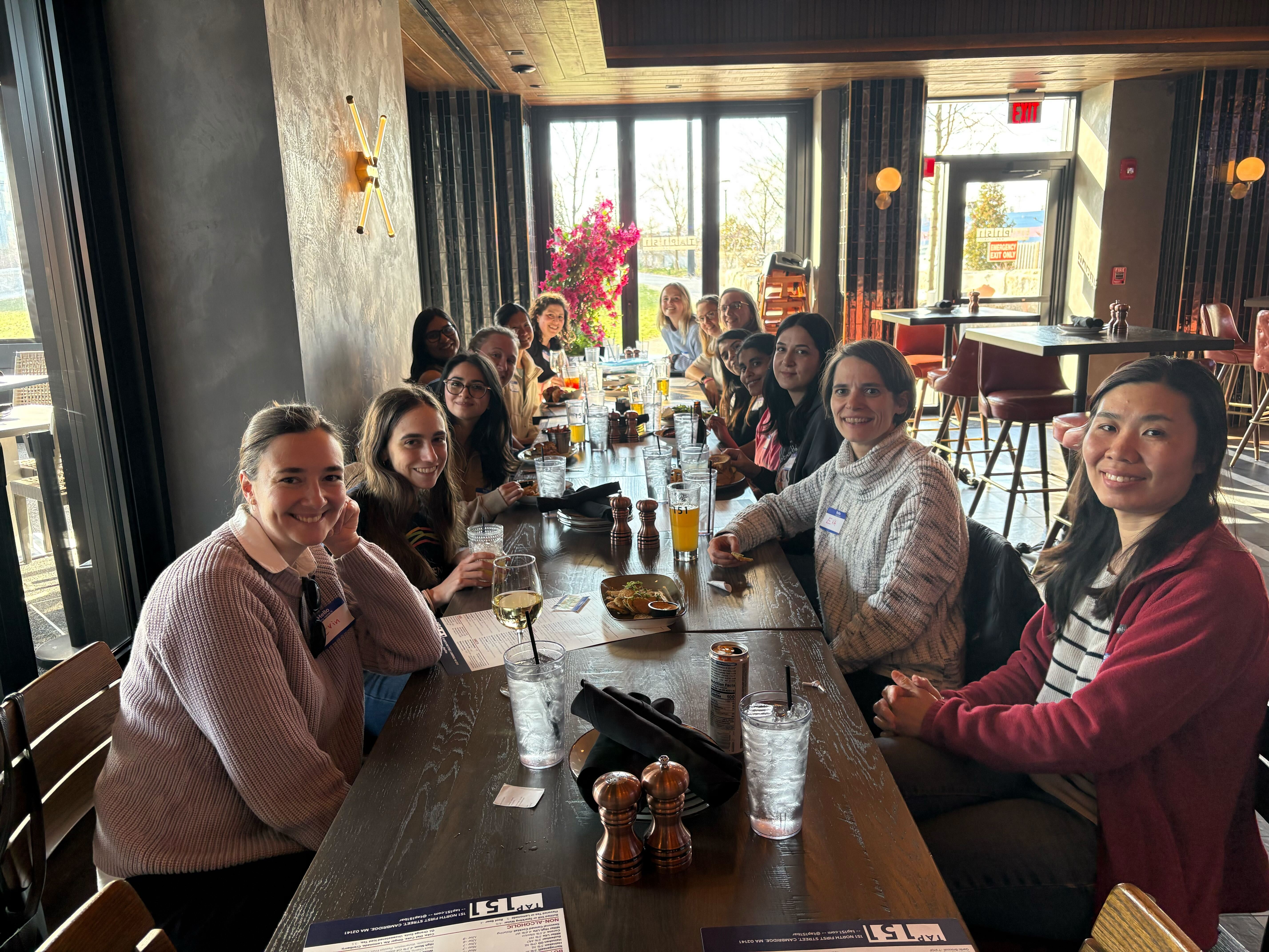 Boston Women in Bioinformatics members seated along a long wooden table, smiling at the camera over drinks.