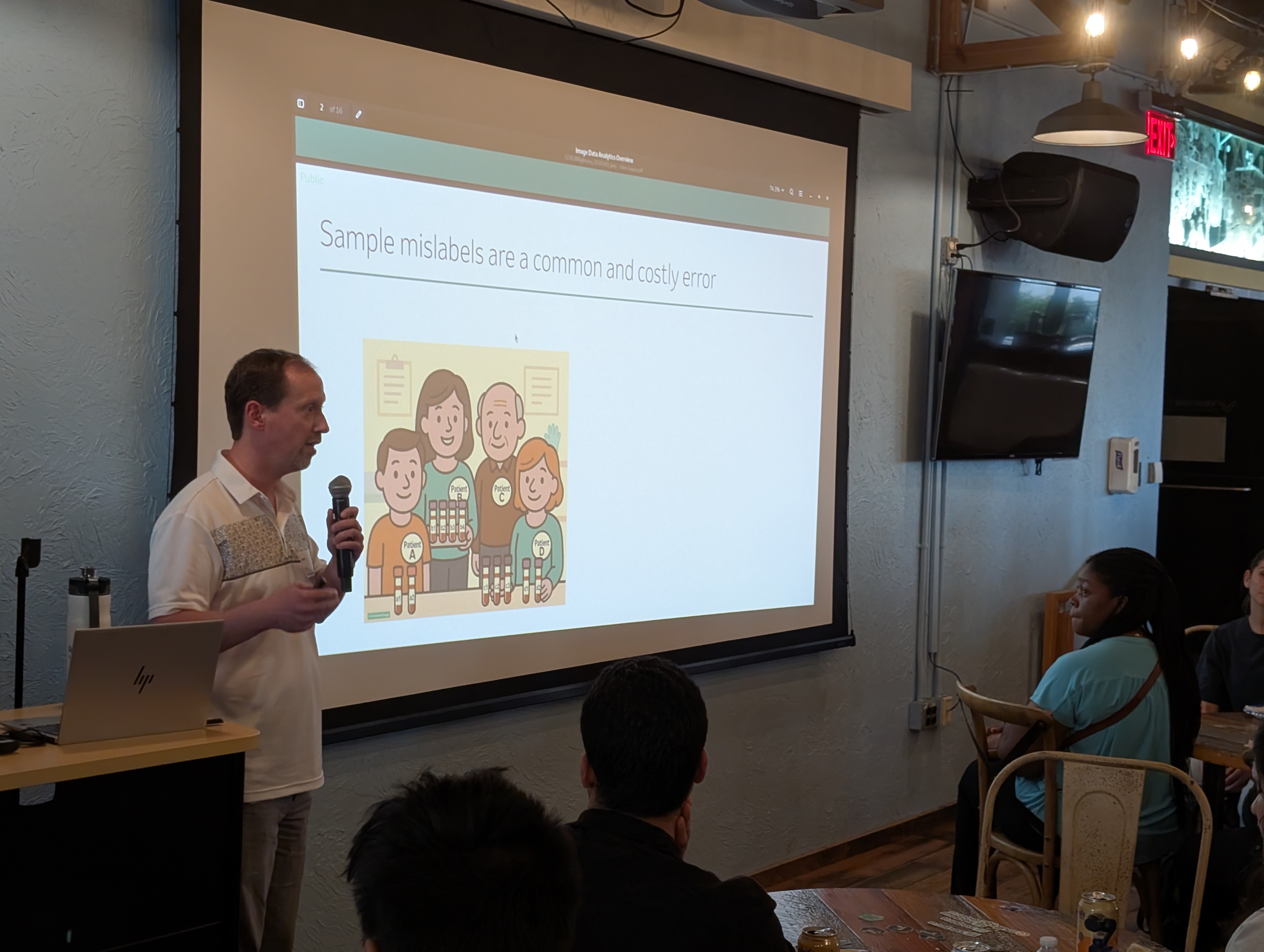 John Gaspar presenting in front of a slide titled "Sample mislabels are a common and costly error." The slide features an illustration of smiling people holding labeled laboratory tubes.