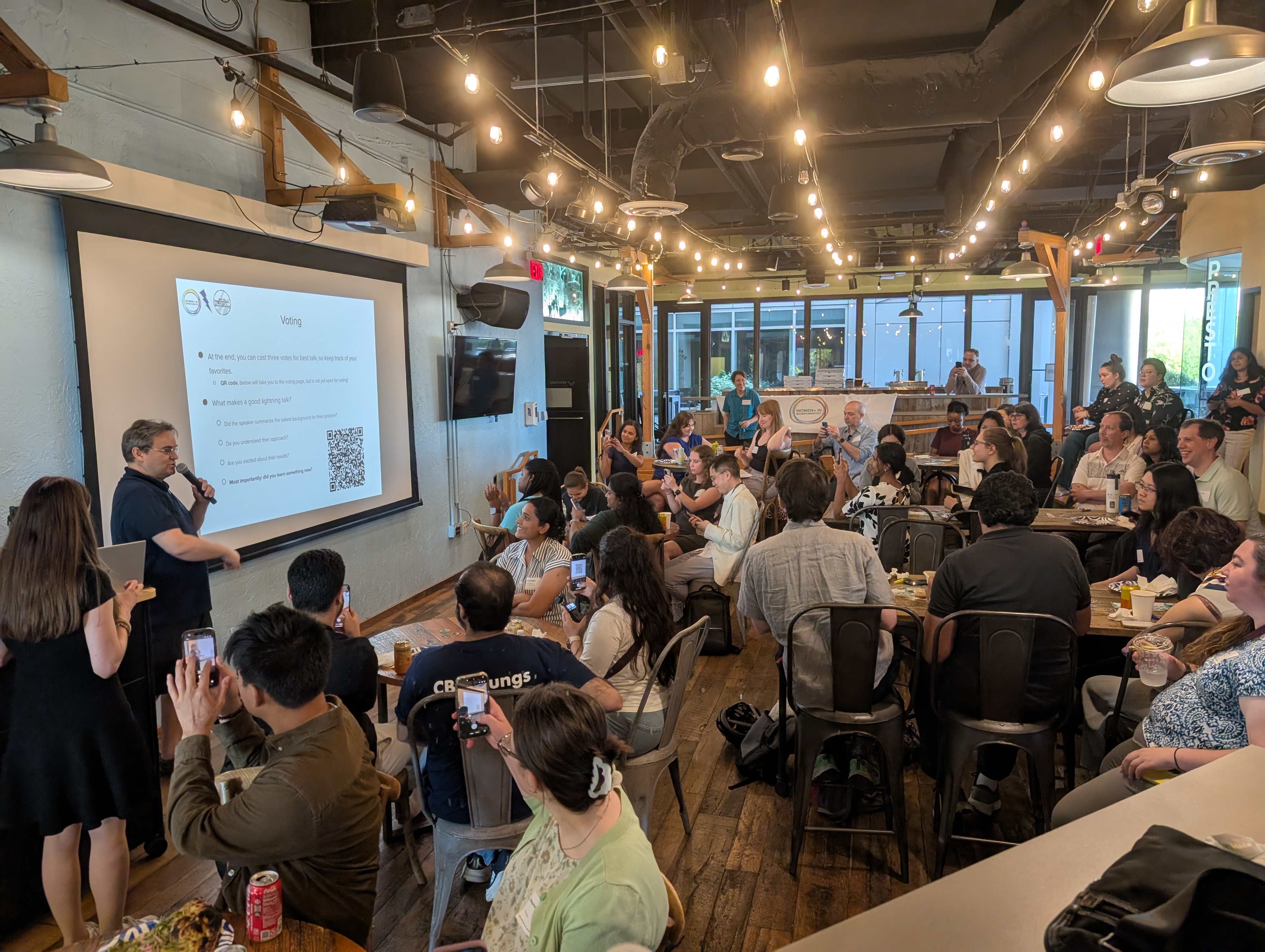 A candid, mid-action photo of an event organizer speaking to an engaged crowd. They are smiling and gesturing with their hands as they explain the voting process for the upcoming lightning talks.